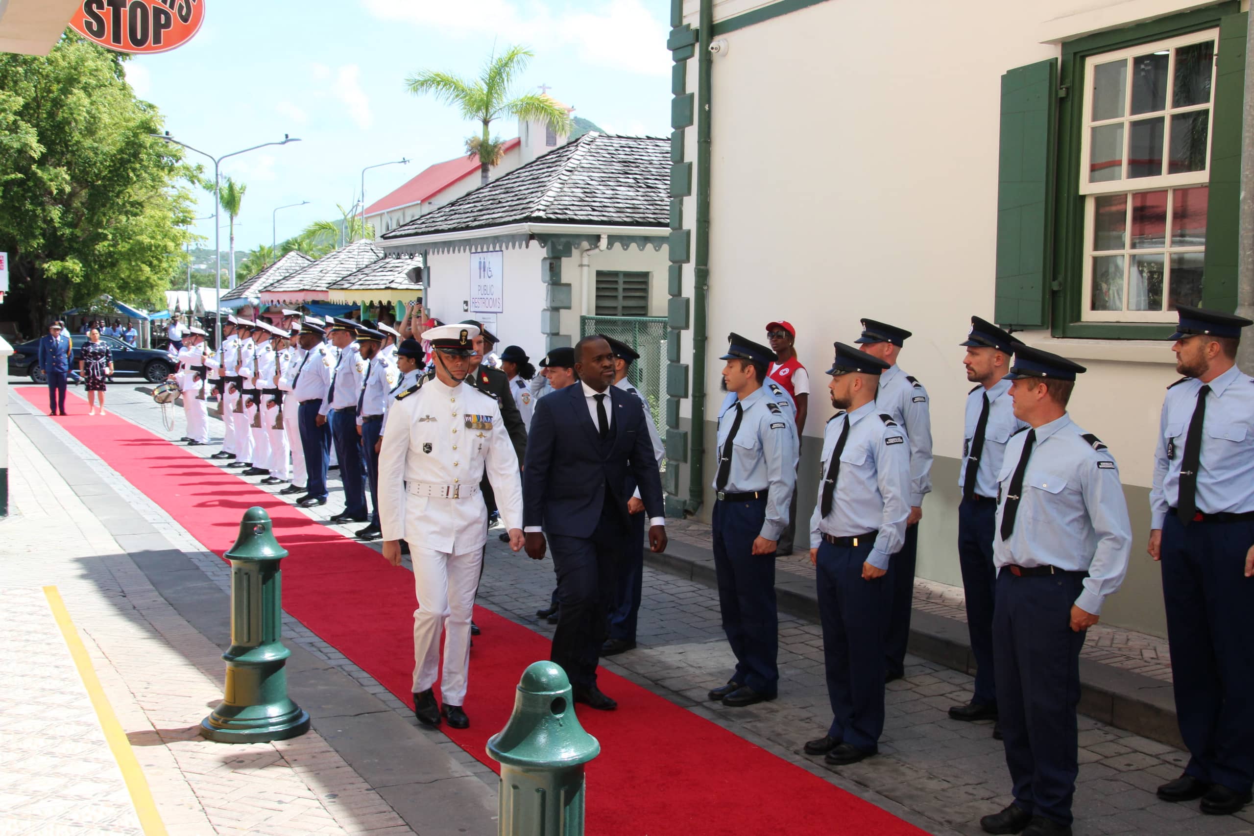 De Gouverneur van Sint Maarten en de adjudant lopen over een rode loper langs een erewacht naar de ingang van het Parlementsgebouw.